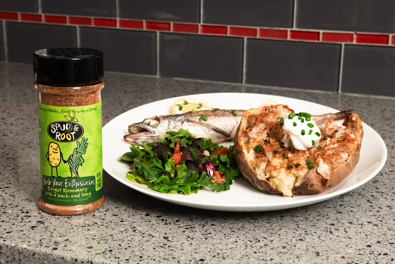 Herb Your Enthusiasm jar on a kitchen counter beside a dinner plate with a seasoned baked potato topped with sour cream and chives, a whole fish and a green salad.