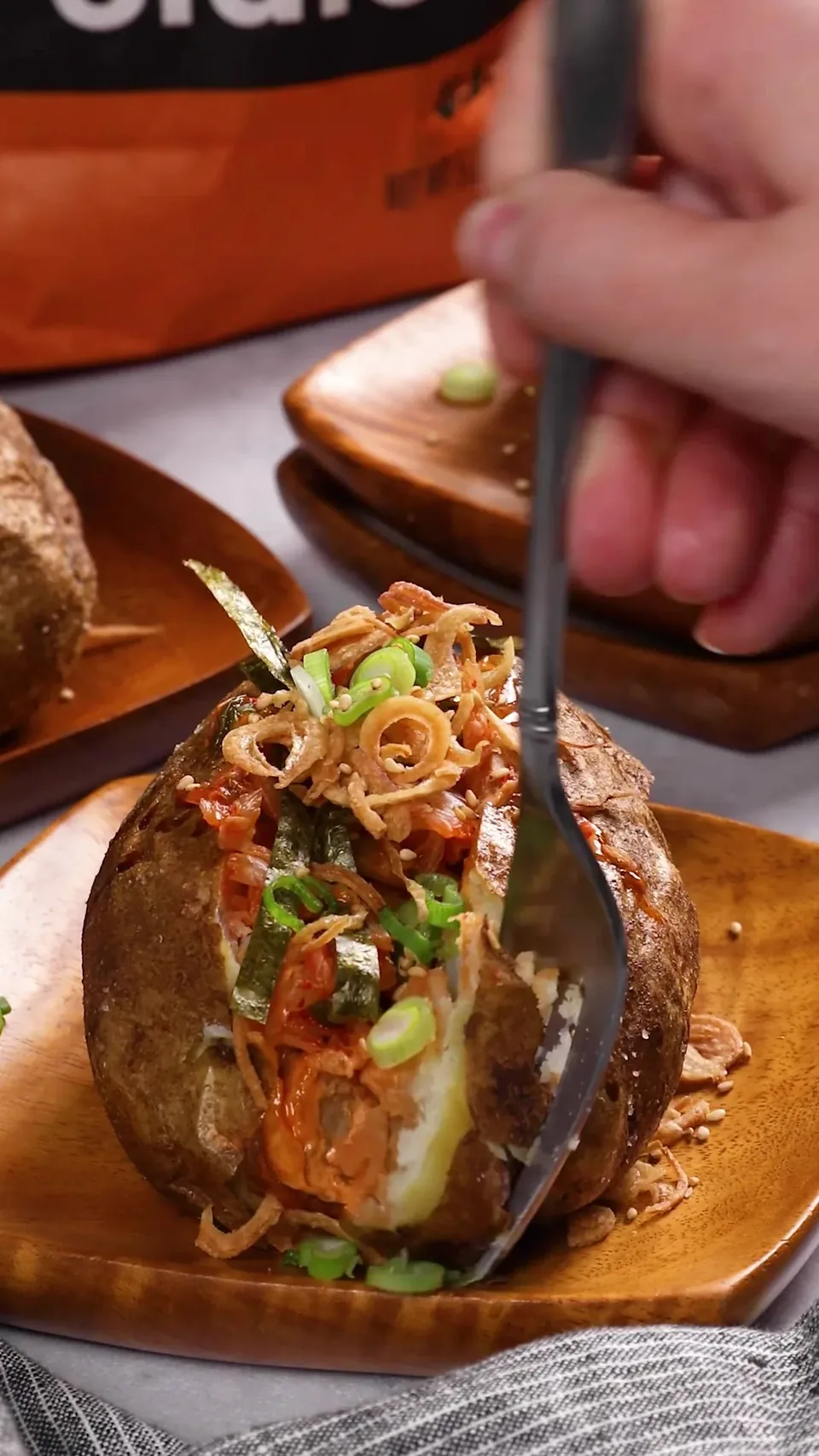 A fork breaking into a baked russet piled with kimchi, spicy mayo, green onions, nori and crispy fried shallots on a wooden dish.