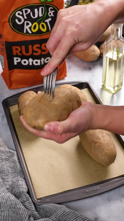 A hand holding a raw russet potato and piercing it with a fork, with a bag of Spud & Root Russet Potatoes behind.