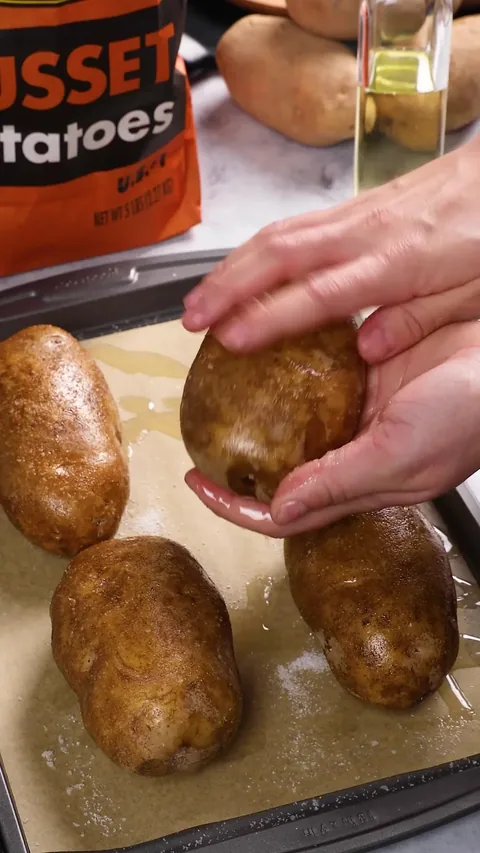 Hands turning an oiled potato on the baking sheet, with salt scattered on the paper.