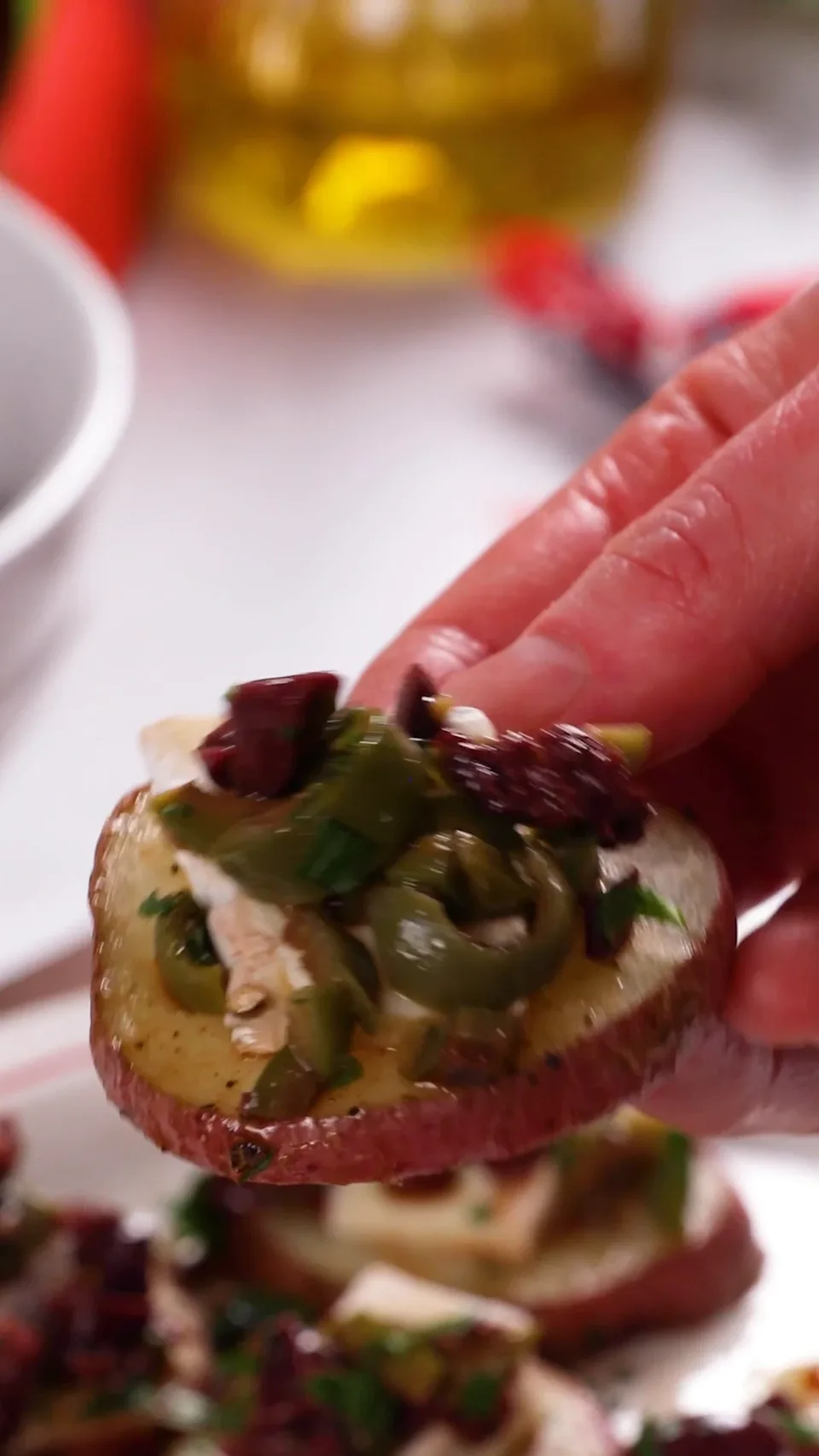 A hand lifting one potato bite, topped with brie and olive tapenade, off the platter.