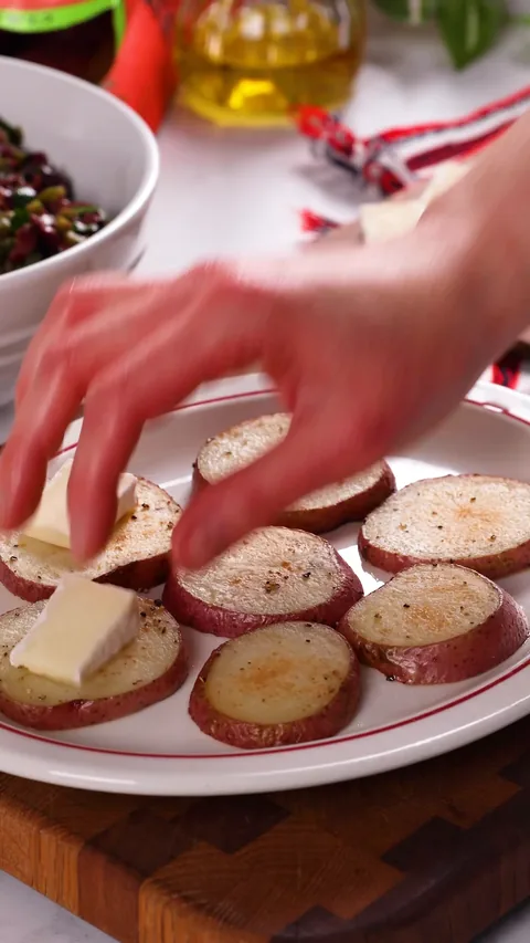 A hand laying a slice of brie on each baked red potato round on a white platter.