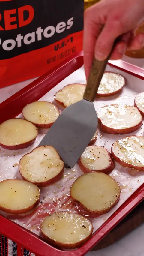 A metal spatula turning over a red potato round on a lined baking sheet.