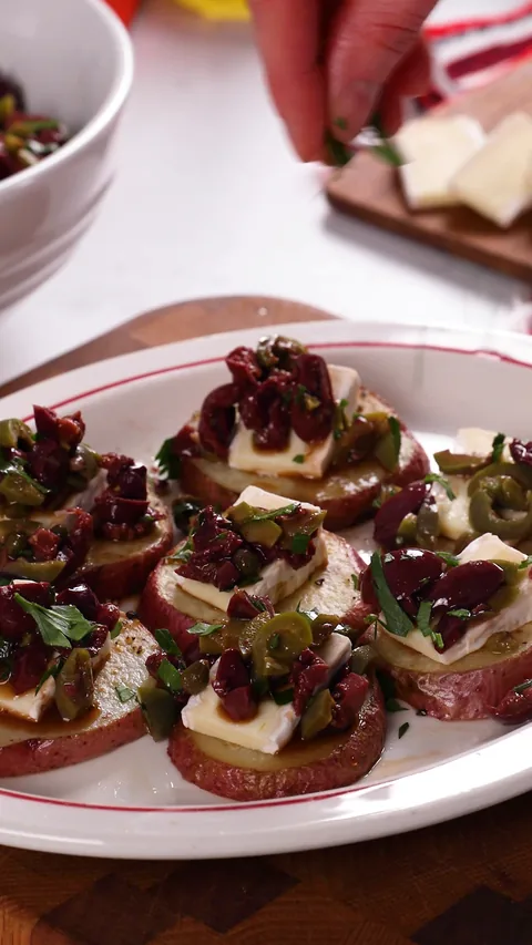 A hand scattering parsley over a platter of potato rounds topped with brie and olive tapenade.