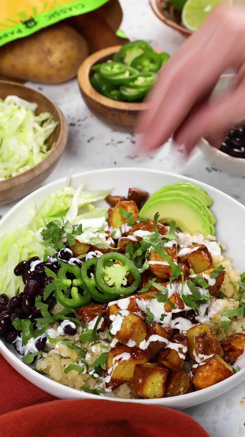 A hand scattering chopped cilantro over a burrito bowl of chipotle-glazed Gold potatoes, black beans, jalapeño rounds, avocado, lettuce and crema on cauliflower rice.