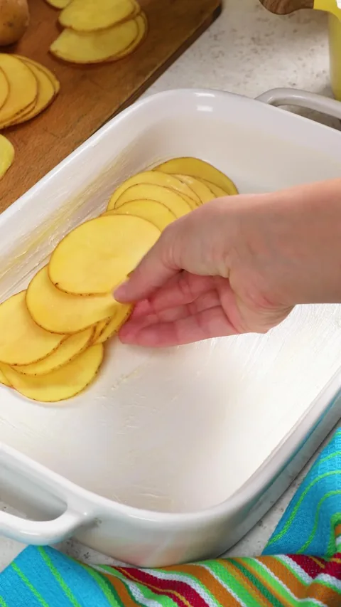 A hand laying thin slices of Gold potato in overlapping rows in a white baking dish.