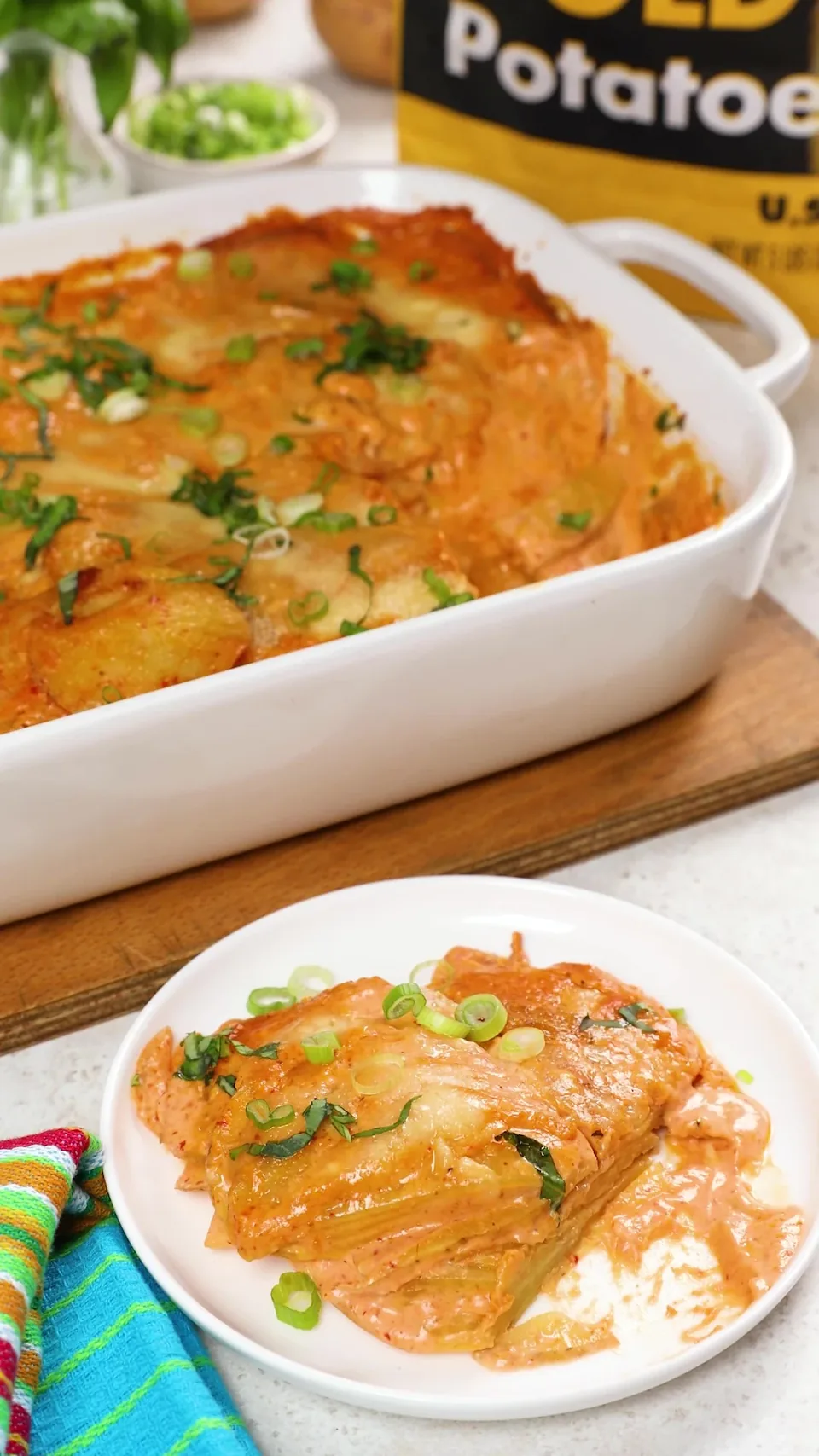 The baked dish of coconut curry scalloped potatoes beside a plate with a serving on it, in front of a bag of Spud & Root Gold Potatoes.