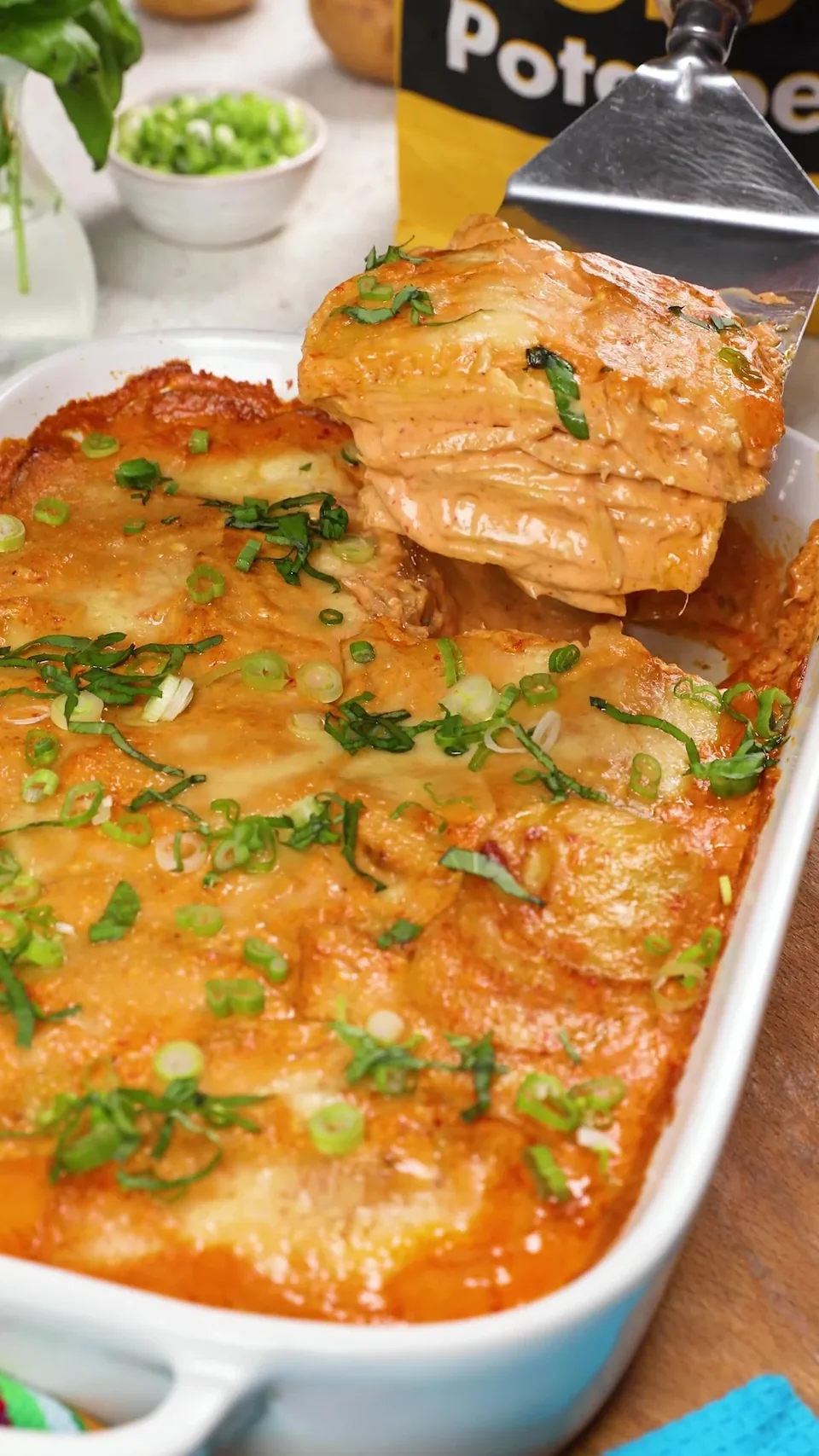 A spatula lifting a layered square of coconut curry scalloped potatoes out of a white baking dish, the top golden and scattered with green onions and basil.