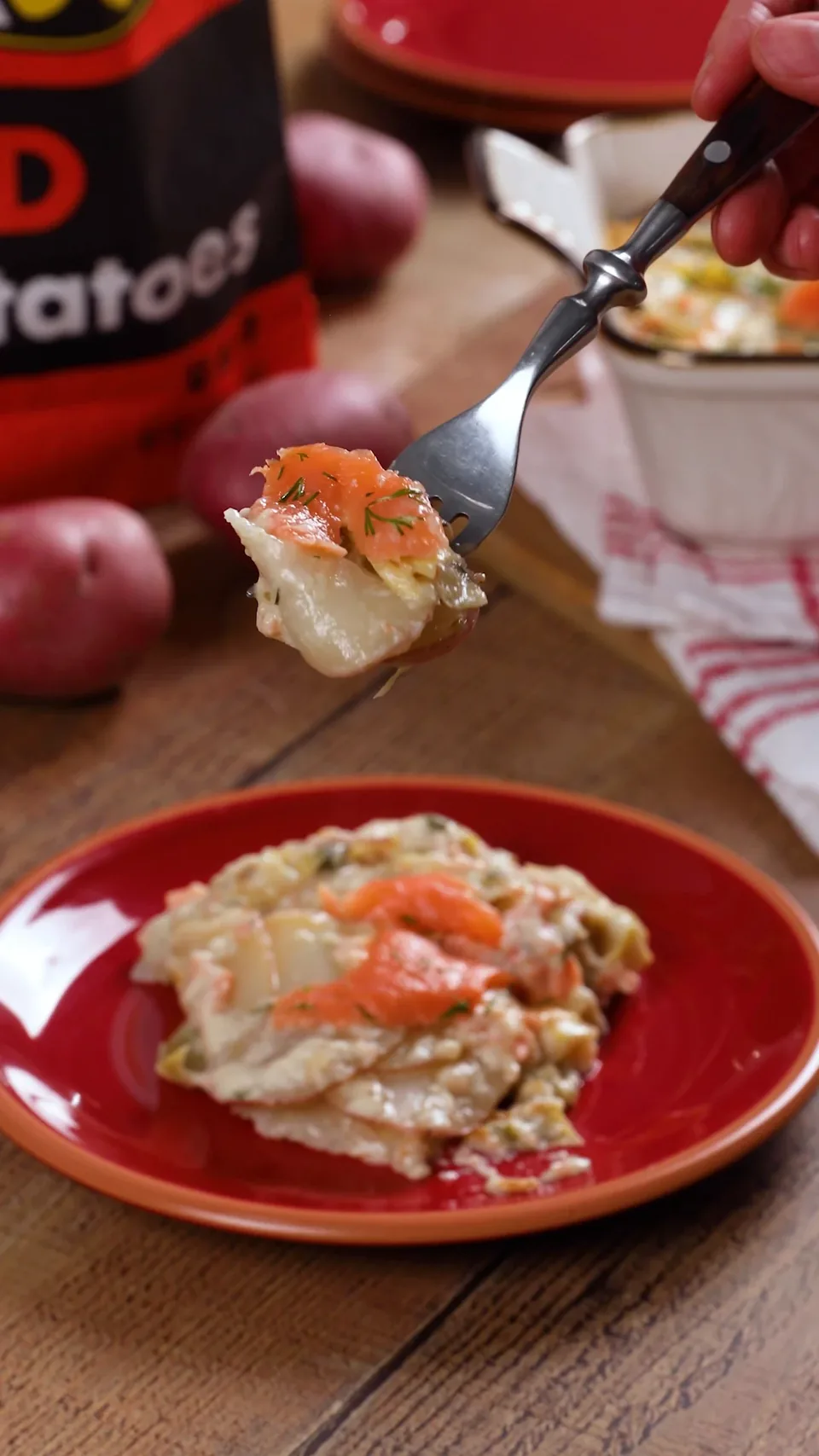 A fork lifting a bite of potato gratin with smoked salmon and dill above a red plate.