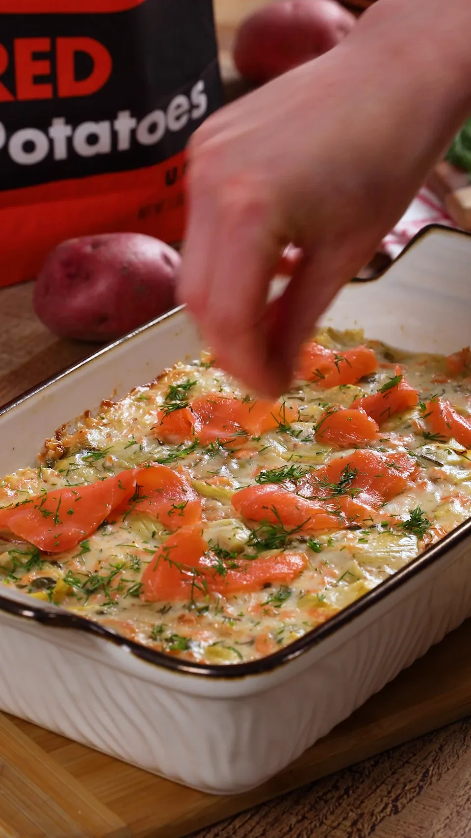 Fresh dill falling from a hand onto a baking dish of potato gratin topped with ribbons of smoked salmon.