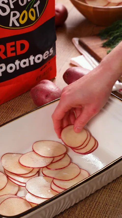 A hand laying thin red potato slices in overlapping rows in a white baking dish.