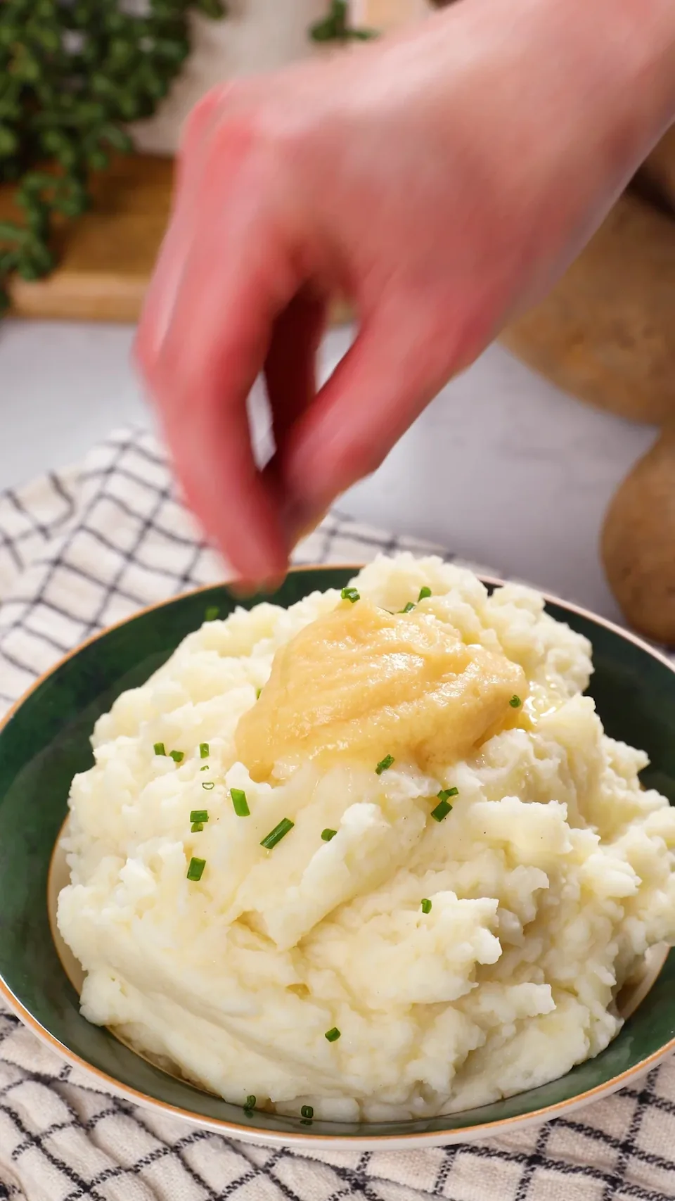 A bowl of mashed potatoes topped with a spoonful of white miso butter and sprinkled with chopped chives, with a hand just above it.