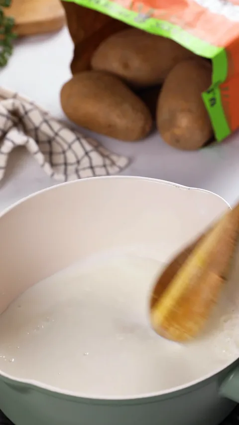A wooden spoon stirring milk in a small saucepan, with a bag of Russet potatoes behind it.