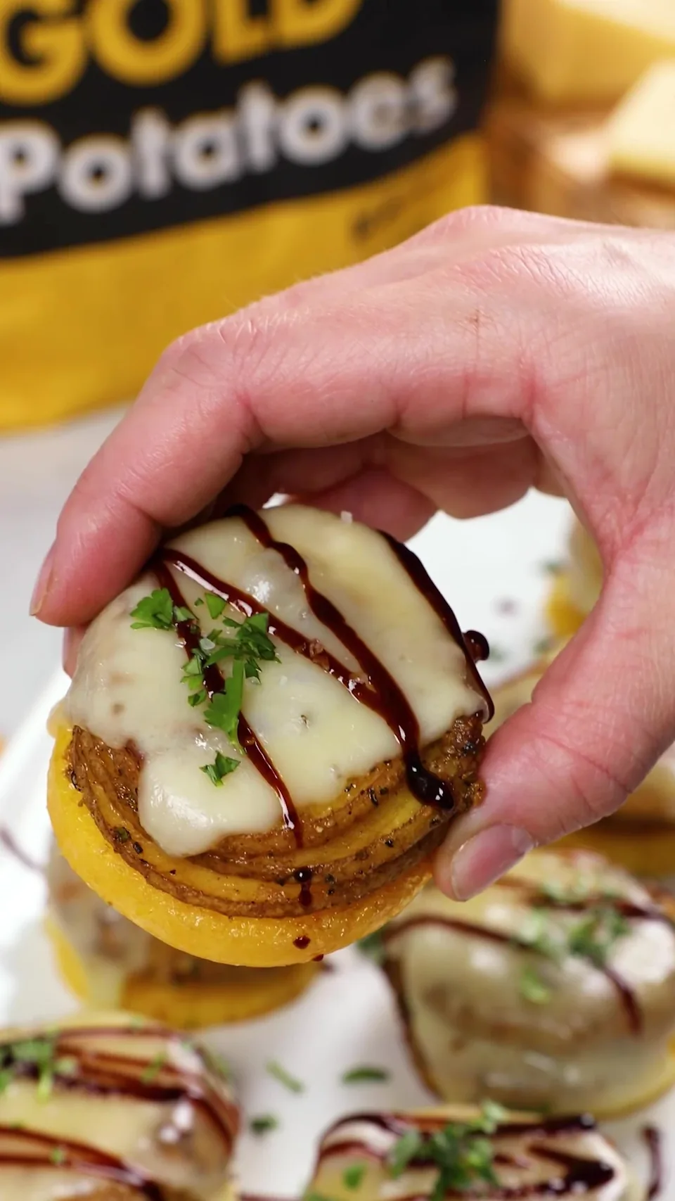 A hand holding up one medallion, dripping with fontina and balsamic glaze, in front of a bag of Spud & Root Gold Potatoes.
