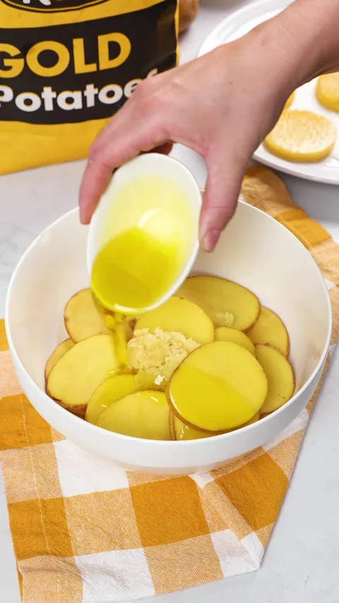 Olive oil being poured over thin potato slices and garlic in a white bowl.