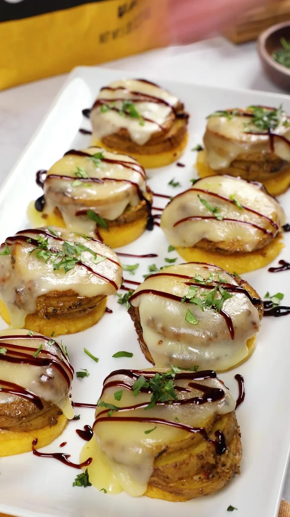 A white platter of polenta and potato medallions, each under melted fontina with a zigzag of balsamic glaze and chopped parsley, with a bag of Spud & Root Gold Potatoes behind.