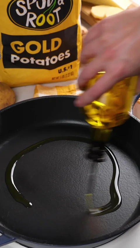 Olive oil being poured into a black cast-iron skillet, with a bag of Spud & Root Gold Potatoes behind.