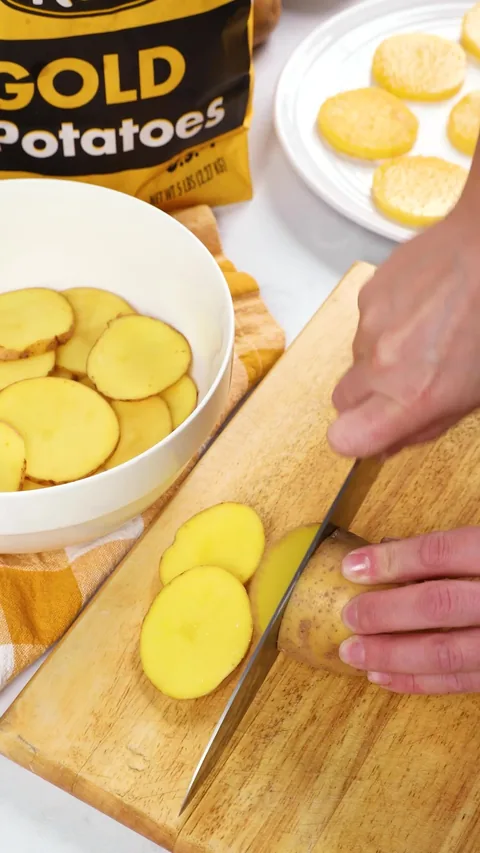 A knife slicing a Gold potato into thin rounds on a wooden board, with a bowl of slices beside it.