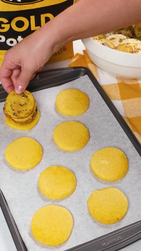 A hand placing a stack of seasoned potato slices on a fried polenta round, on a lined baking sheet.