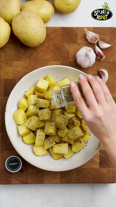 Dried oregano shaken over the seasoned potato cubes in the bowl.