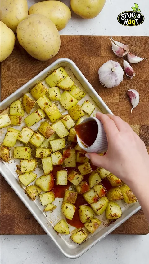 A small cup pouring dark red sauce over the roasted potato cubes on the baking sheet.