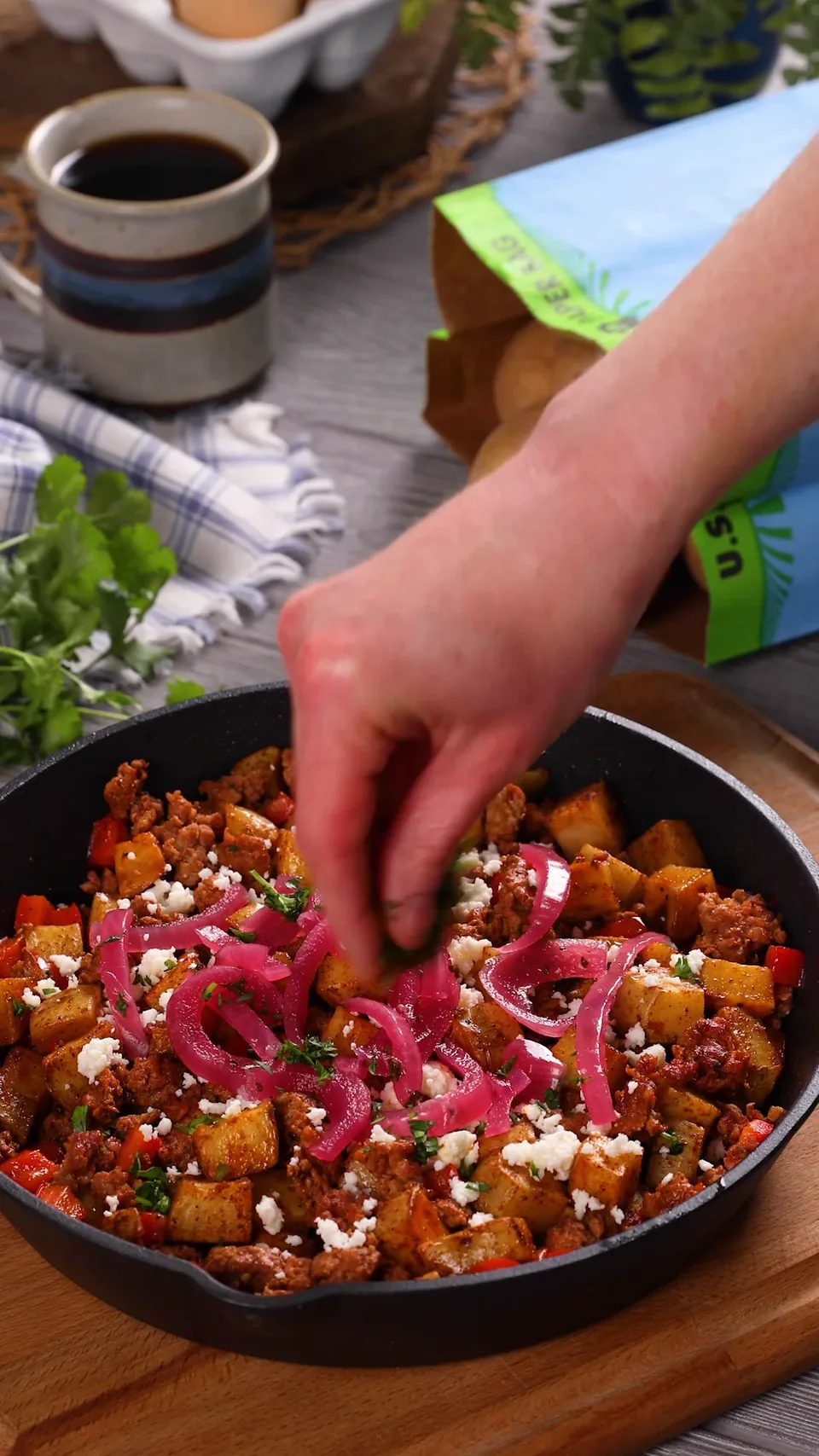 A hand scattering cilantro over a cast-iron skillet of golden potatoes, chorizo and red pepper, topped with pickled red onions and queso fresco.
