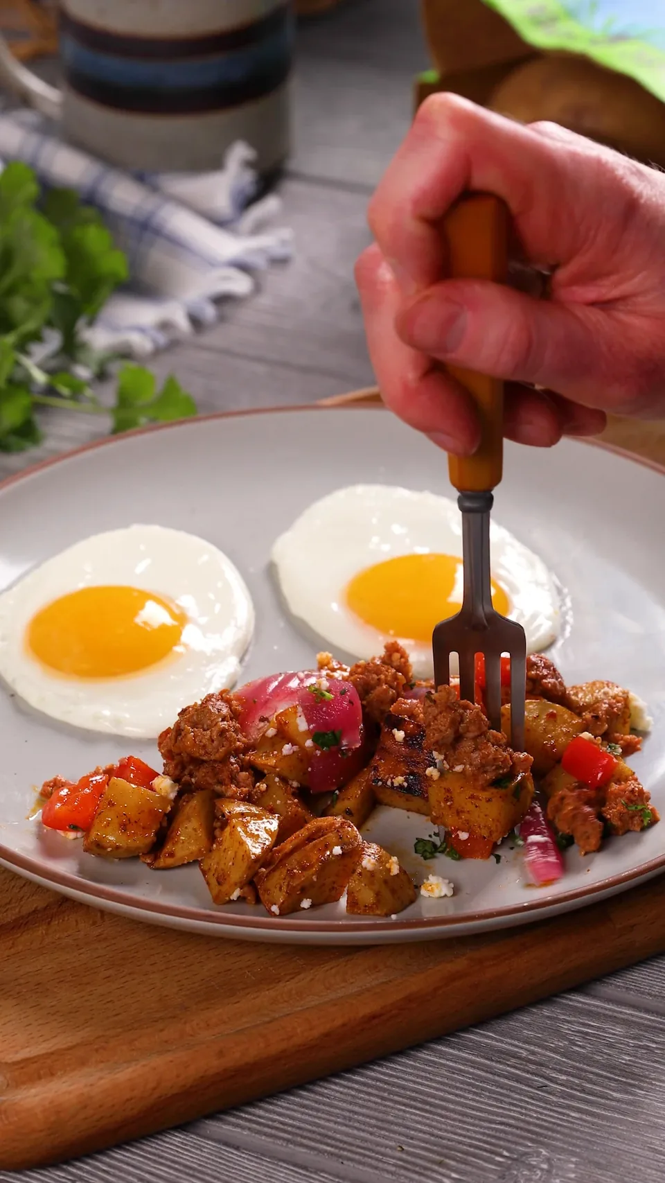 A fork digging into potatoes and chorizo on a plate beside two fried eggs.