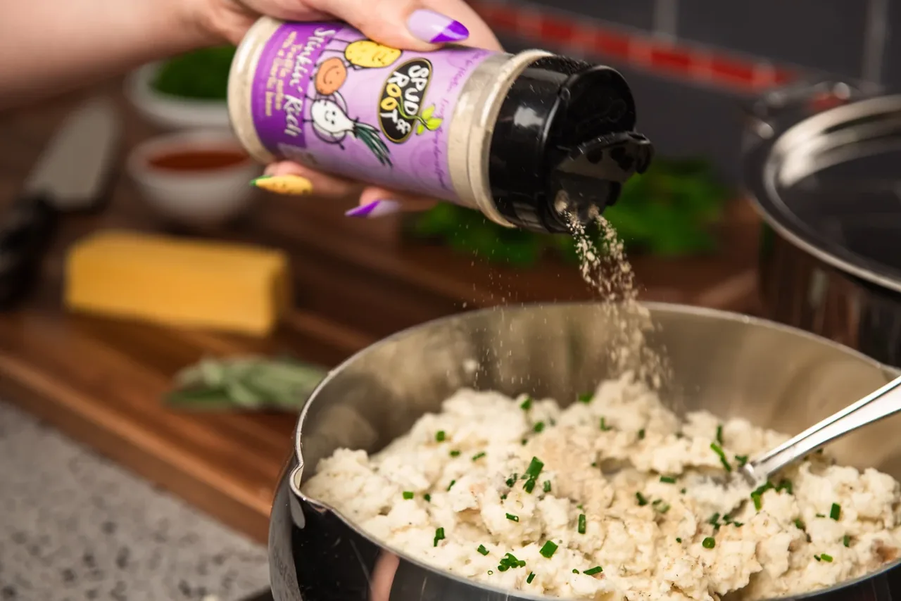A hand shaking Stinkin' Rich over a pot of mashed potatoes with chives, beside a block of cheddar and fresh herbs.